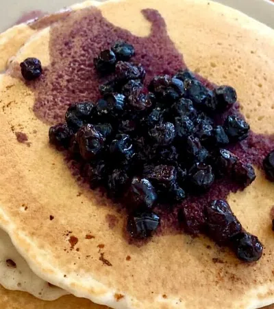 Stack of golden brown pancakes topped with juicy cooked blueberries and blueberry syrup on a plate.