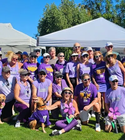 Group of people wearing purple shirts posing outdoors with two dogs under white tents on green grass