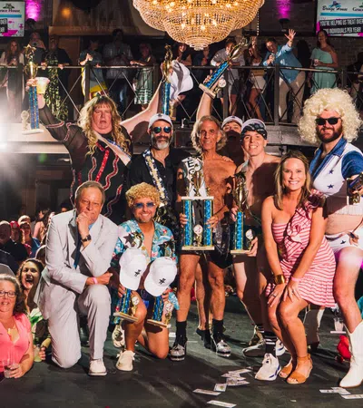 Group of people in colorful costumes holding trophies on stage with cheering audience and chandelier overhead