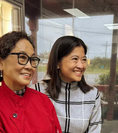 Two women smiling inside a bakery, one wearing a red coat and glasses, the other in a gray plaid sweater.