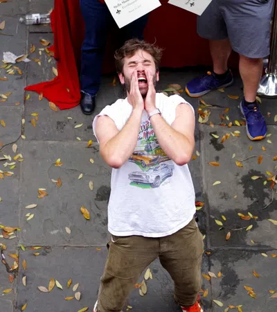 Man inside red ropes shouting or calling out while onlookers stand outside on a leaf-strewn pavement.