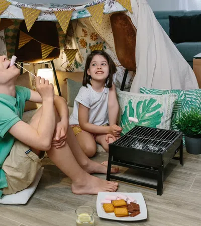 Two children enjoying indoor camping with a blanket tent and roasting marshmallows on a small grill.