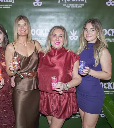 Four women posing with Mockly non-alcoholic beverage cans in front of a branded green backdrop at an event.