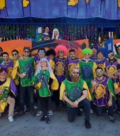 Group of people wearing colorful Mardi Gras costumes and wigs posing in front of festive decorations outdoors.