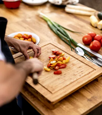 Person chopping red and yellow cherry tomatoes on a wooden cutting board with fresh vegetables nearby