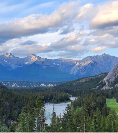 River winding through dense forest with towering mountains and cloudy blue sky in the background