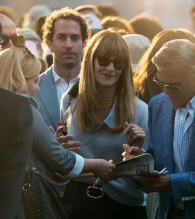 Celebrity signing autographs for fans outdoors in stylish attire during daytime event with crowd around.