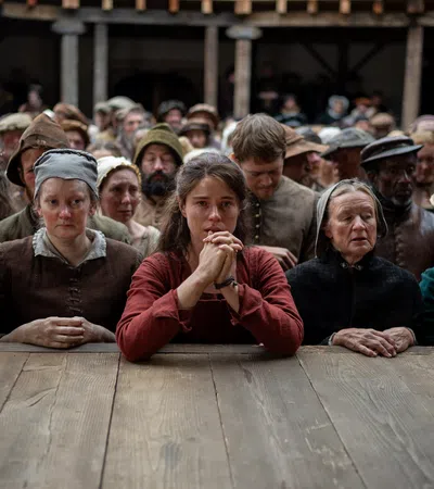 Crowd of somber medieval villagers in simple clothing gathered around a wooden table in a rustic hall.