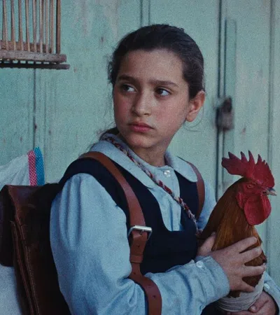 Young girl holding a rooster while sitting outdoors against a weathered wooden wall.