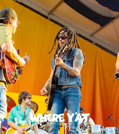 Musicians perform energetically on stage with guitars, tambourine, and drums in front of a yellow backdrop.