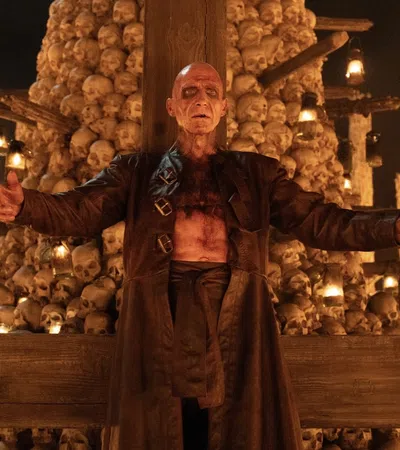 A bald man with scars stands in front of a towering pile of skulls and lit lanterns, spreading his arms wide.