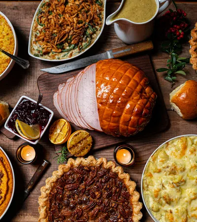 Thanksgiving feast with glazed ham, pecan pie, pumpkin pie, mashed potatoes, stuffing, rolls, and gravy on wooden table.