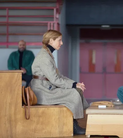 Two women seated on wooden benches facing each other in a stark room, engaged in serious conversation.
