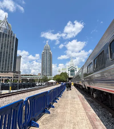 Silver train at platform with blue seats and city skyscrapers under a partly cloudy sky.