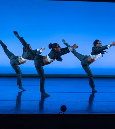 Three dancers in gray suits perform a high leg balance on a blue-lit stage in unison.