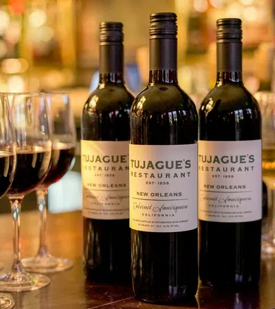 Three bottles of Tujague's Restaurant red wine with six filled wine glasses on a wooden bar counter.