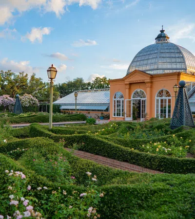 Historic glass-domed conservatory with manicured garden and hedges under a blue sky with clouds.