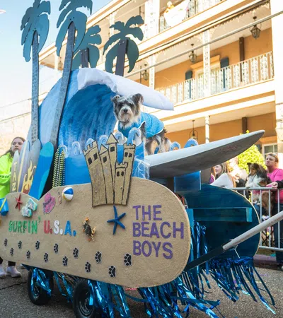 Dog in blue sweater on a beach-themed parade float with surfboard and palm tree decorations during street event