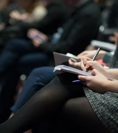 Participants engaged in a conference, taking notes in notebooks during a presentation.