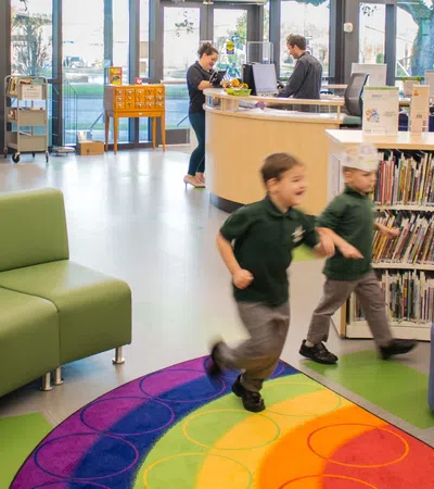 Children running on a colorful carpet in a modern library with bookshelves, seating, and large windows in the background
