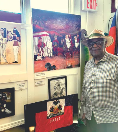 Elderly man in a straw hat stands next to Haitian flag and cultural paintings inside a bright gallery space.