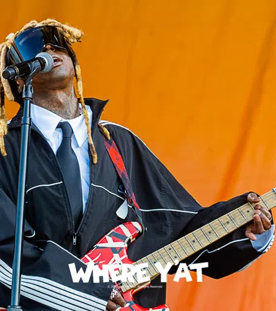 Musician performing on stage, wearing a black jacket and playing a vibrant guitar.