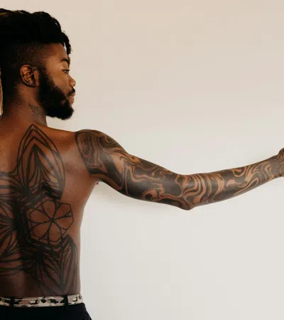 Man with detailed geometric tattoos covering his back and arm, pointing to the side against a plain background.