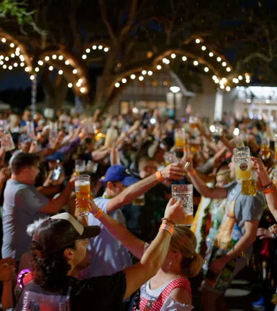 Crowd enjoying outdoor night event raising glasses of beer under string lights and large tree.