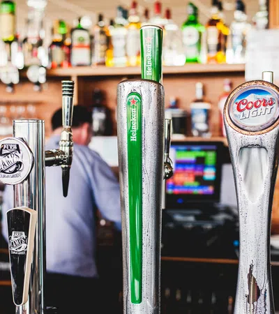 Close-up of multiple beer taps including Heineken, Coors Light, and others in a bar setting with blurred bartender.