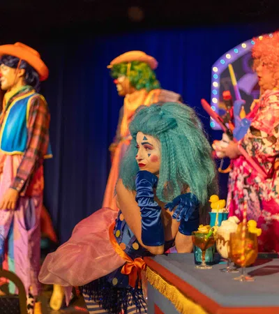 Colorful clowns in vibrant costumes perform on stage with drinks on a table under theatrical lighting.