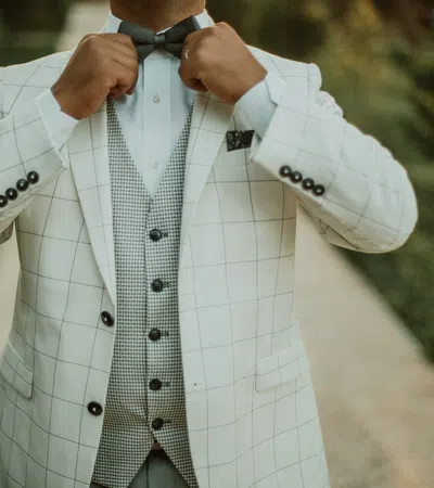 Man adjusting bow tie wearing a white checkered suit jacket and houndstooth vest outdoors on pathway.