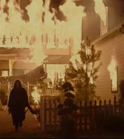 Person walks towards a large house engulfed in flames at night, amidst a white picket fence and trees.