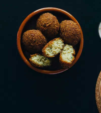 Bowl of golden falafel balls with cut pieces showing inside, served with creamy dip and hummus on black background