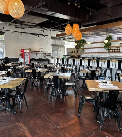 Modern restaurant interior with wooden tables, black metal chairs, pendant lights, and a long bar counter.