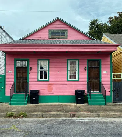 Pink wooden house with green trim, two doors, windows, and trash bins on a city sidewalk in daytime.