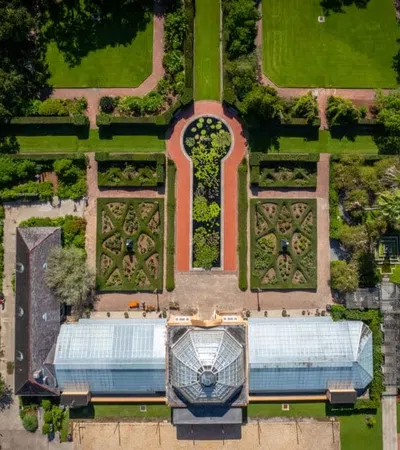 Aerial view of a symmetrical botanical garden with pathways, green lawns, manicured hedges, and a glass conservatory.