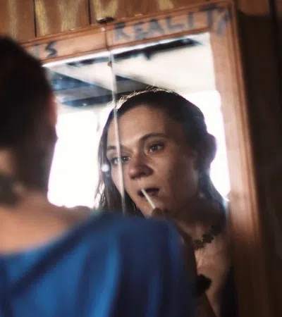 Woman applying lipstick while looking into a vintage wooden mirror in a cozy room.