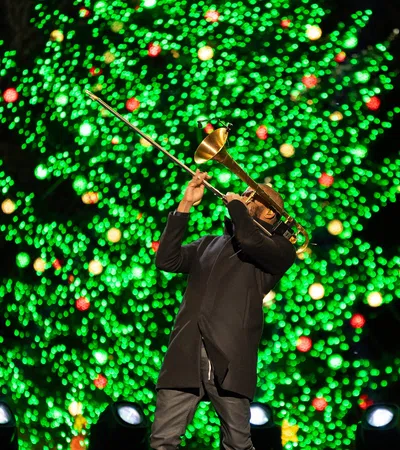 Musician playing trombone in front of a brightly lit Christmas tree with green and red lights at night.