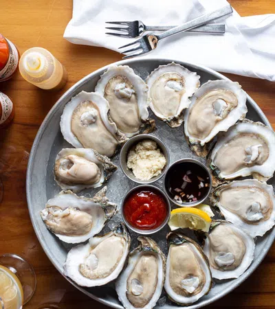 Plate of fresh oysters on the half shell with cocktail sauce, lemon, and condiments on a wooden table with drinks
