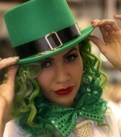 Woman with green curly hair wearing a green top hat and matching sequined bow tie, posing indoors.