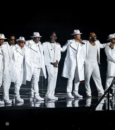 Nine men in coordinated white suits and hats performing together on stage with microphones under spotlight.
