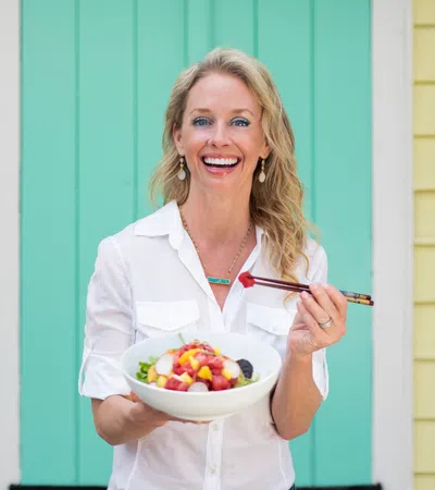 Smiling woman in white shirt holding a bowl of fruit salad with chopsticks in front of pastel yellow and green walls