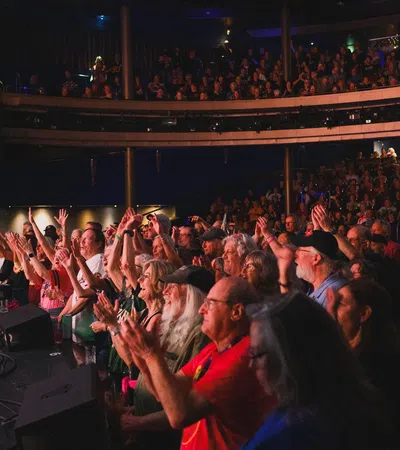 Audience cheering and applauding at an indoor live concert with a multi-level venue filled with spectators