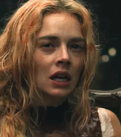 Blonde woman with disheveled hair and distressed expression sitting on an ornate wooden chair in dim lighting.