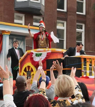 Crowd waves at a parade float featuring a man in Roman armor and two men in suits in a city setting.