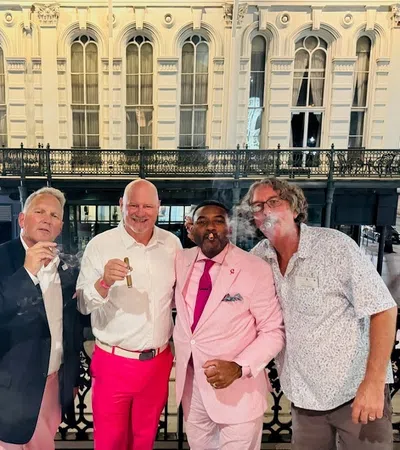 Group of five men dressed smartly enjoying cigars on a balcony with ornate historic building in background
