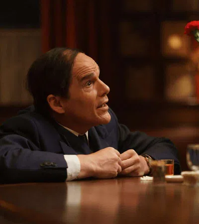 Man in suit sitting at a bar counter with whiskey and red flowers in a dimly lit setting.
