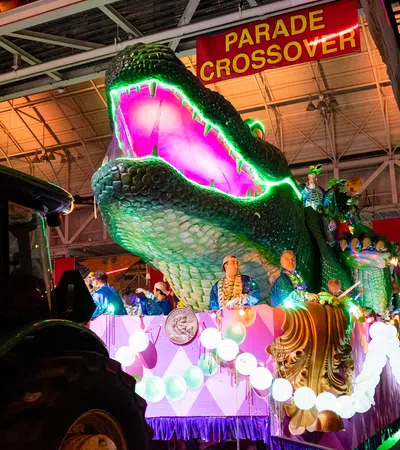 Large illuminated alligator float with purple and green lights at a nighttime parade under a parade crossover sign.