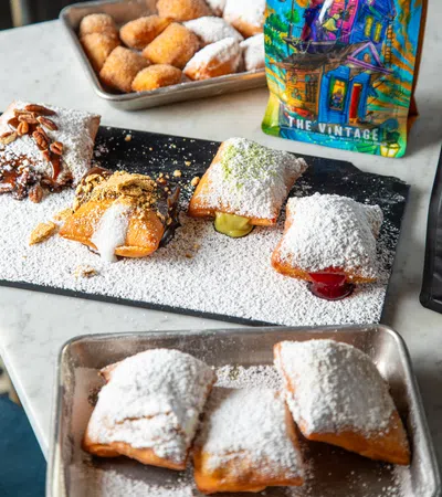 A vibrant display of powdered beignets and desserts at a cafe with colorful artwork in the background.