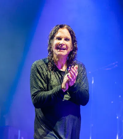 Rock musician with wet hair and black nail polish clapping on stage under blue spotlight with microphone nearby.